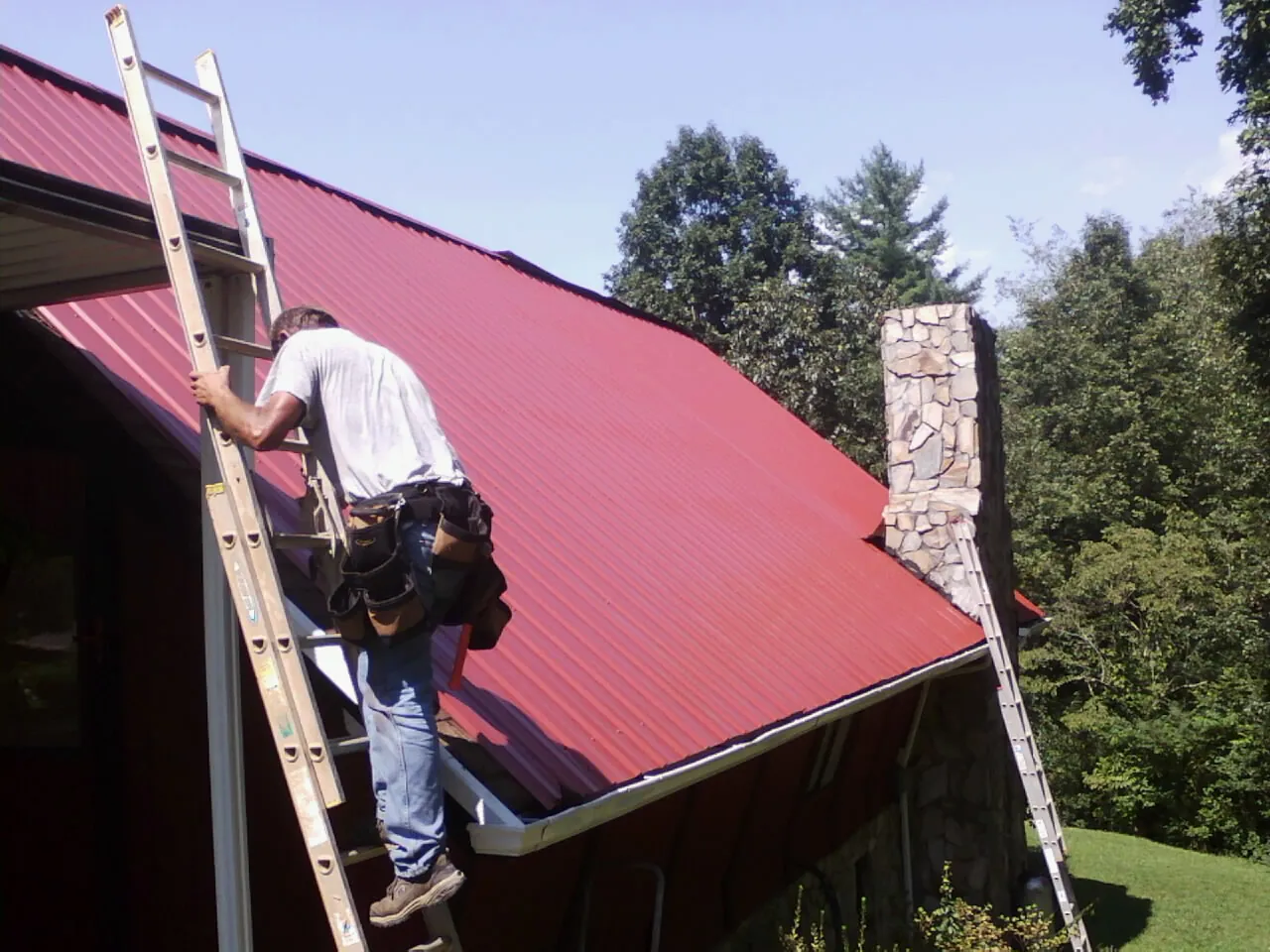 Roofer on a ladder beside a red metal roof and stone chimney — Soffit & Fascia Repair by Roofing Brothers in Flandreau